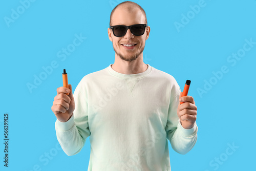 Young man in sunglasses with disposable electronic cigarettes on blue background