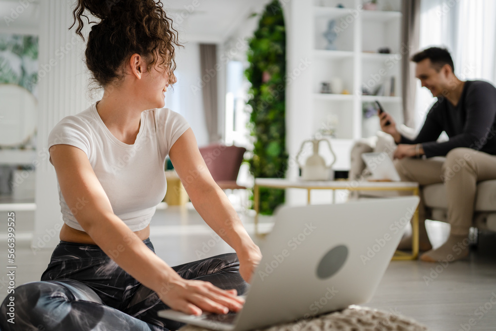 Fototapeta premium Woman young adult caucasian female sitting on the floor at home working on laptop computer browse internet online while her husband or boyfriend is sitting in the background leisure family concept