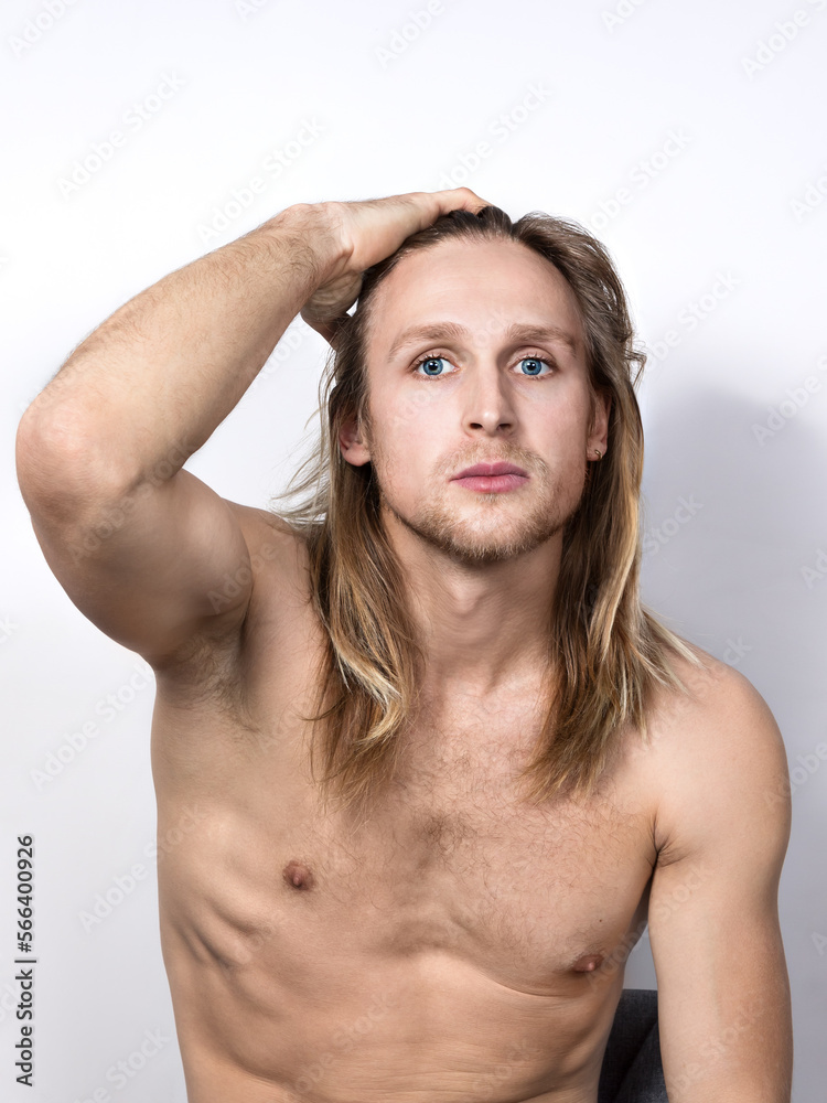 A handsome,seductive, topless young blond man with blue eyes sits on a chair,touching his long hair with his hand, and looks into the camera.Portrait of a real guy to the waist on a white background A handsome,seductive, topless young blond man with blue eyes sits on a chair,touching his long hair with his hand, and looks into the camera.Portrait of a real guy to the waist on a white background