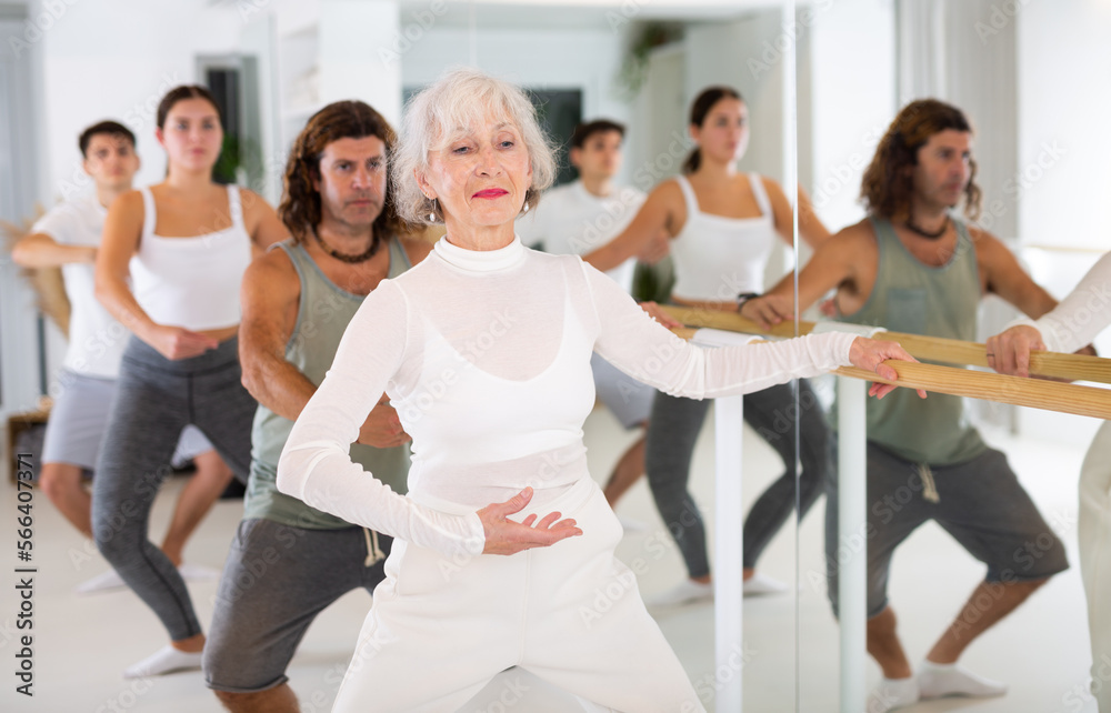 Fototapeta premium Gracile old-aged woman engaging in ballet at ballet barre in training hall during workout session