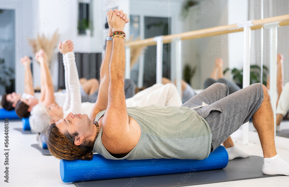Fototapeta premium Diligent middle-aged man practicing pilates with blue roller in training area during pilates classes