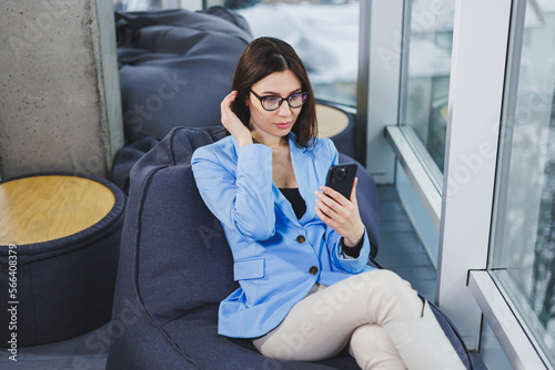 Business young woman in glasses with long dark hair in casual clothes smiling and looking at the phone, browsing the smartphone during a day off in the workspace © Дмитрий Ткачук