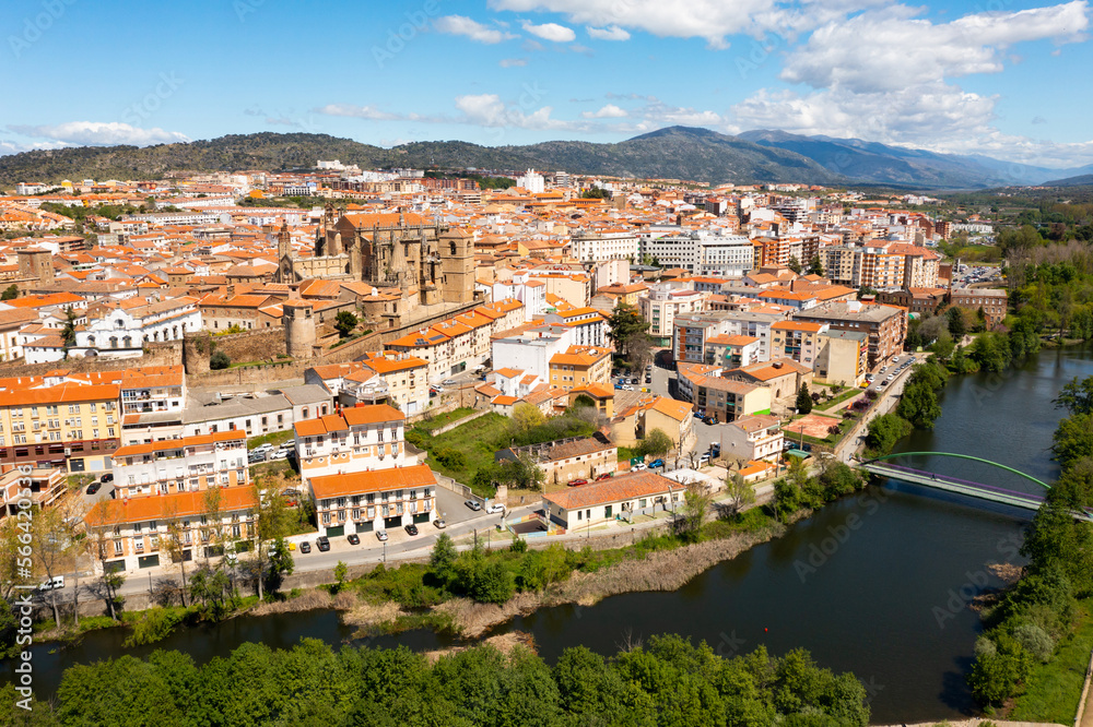 View from drone of residential area of Plasencia city with brownish tiled roofs of houses and ancient gothic roman catholic cathedral on green bank of meandering Jerte river in spring, Caceres, Spain
