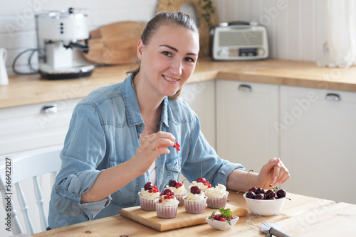 A young woman decorates muffins with white cream and berries in her kitchen.