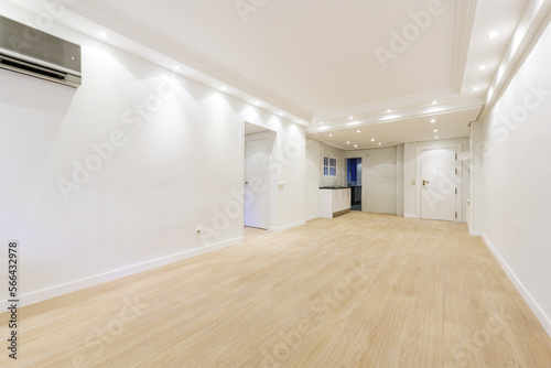 Empty living room with white walls, newly installed laminated floors, kitchen in the background and white access door