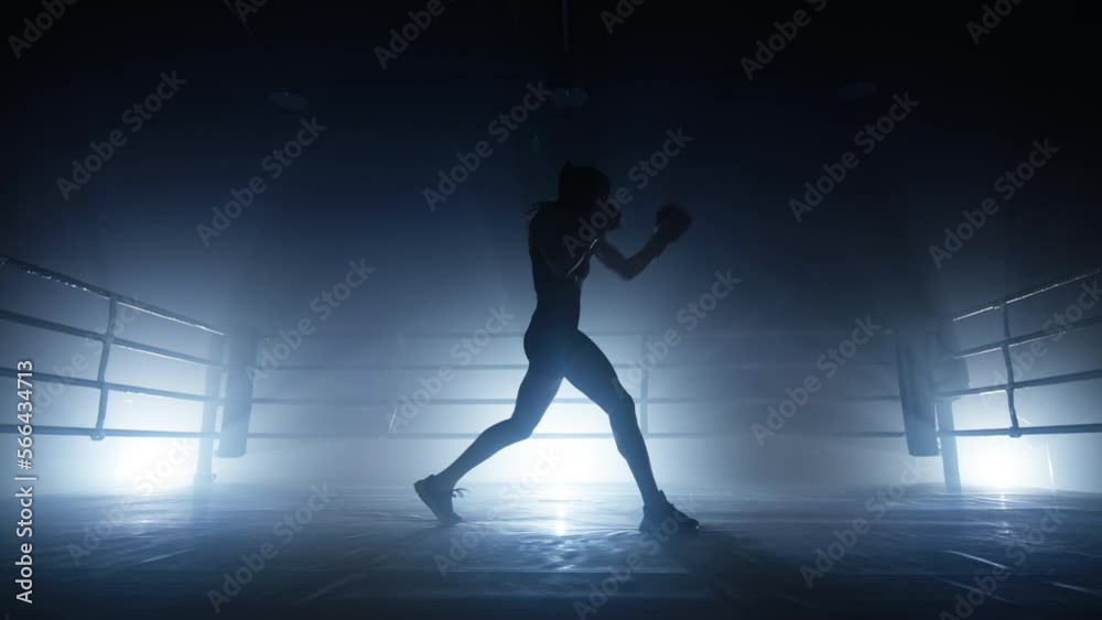 Unrecognized girl having intensive workout at boxing club alone. Close-up view of female athlete kicking and punching in gloves. High quality 4k footage in cold blue purple silver foggy back light