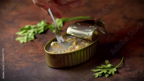 Hand with a fork picking up canned cockles on a dark brown table with parsley. Ready to eat.