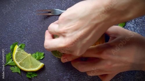 Hands opening a can of sardines with tomato sauce on a dark blue table with parsley. Ready to eat.