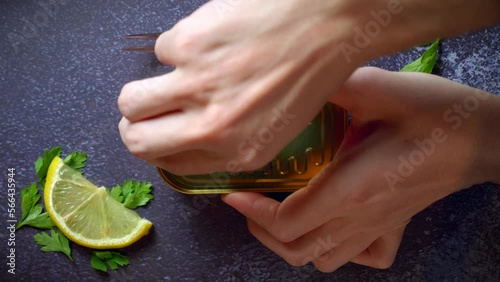 Hands opening a can of sardines on a dark blue table with parsley. Ready to eat.