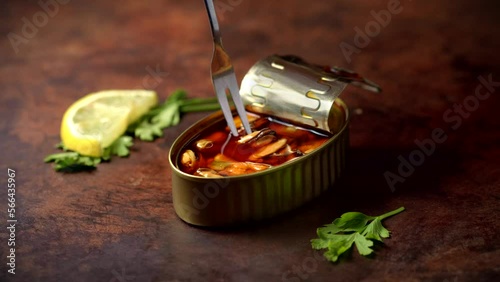 Hand with a fork picking up canned mussels on a dark brown table with lemon and parsley. Ready to eat.