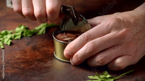 Hands opening canned octopus with a old can opener on a dark brown table with parsley. Ready to eat.