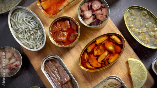 Preserve cans with different products, fish and seafood, natural or pickled, open on a dark table with a wooden cutting board and lemon. Ready to eat.