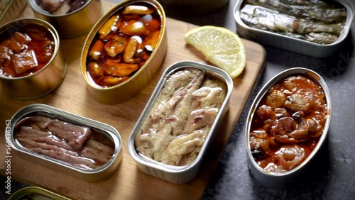 Preserve cans with different products, fish and seafood, natural or pickled, open on a dark table with a wooden cutting board and lemon. Ready to eat.