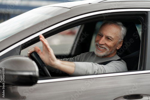 Cheerful gray-haired male driver, smiling and waving with hand while driving newly bought luxury car