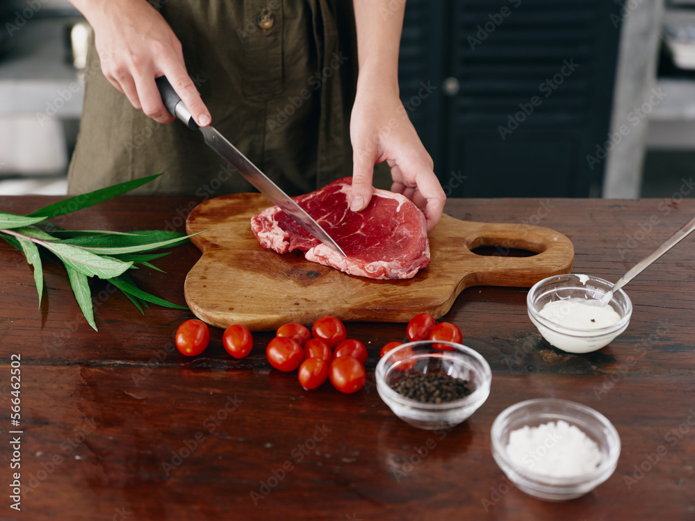 Woman with knife in hand cutting steak meat for frying in kitchen with salt pepper and other spices on table, red cherry tomatoes and herbs, dinner preparation.