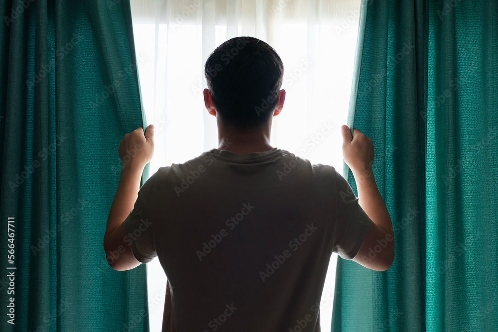 Man stands at window and opens turquoise curtains rear view.