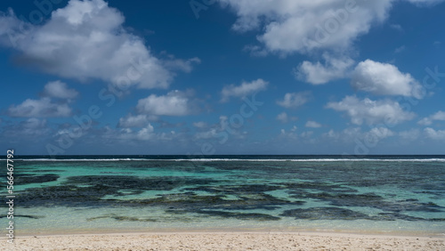 Corals are visible through the clear water of the turquoise ocean. The waves are foaming over the reef in the distance. Picturesque clouds in the blue sky. Sandy beach in the foreground. Seychelles.