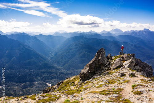 Adventurous athletic female hiker standing on top of a rugged mountain in the Pacific Northwest with jagged mountains in the background.

