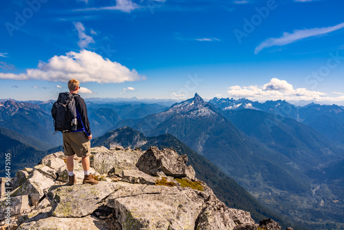 Adventurous athletic male hiker standing on top of a rugged mountain in the Pacific Northwest with jagged mountains in the background. 