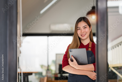Smiling young woman in apron holding tablet standing at cafeteria door entrance. Successful small business owner.