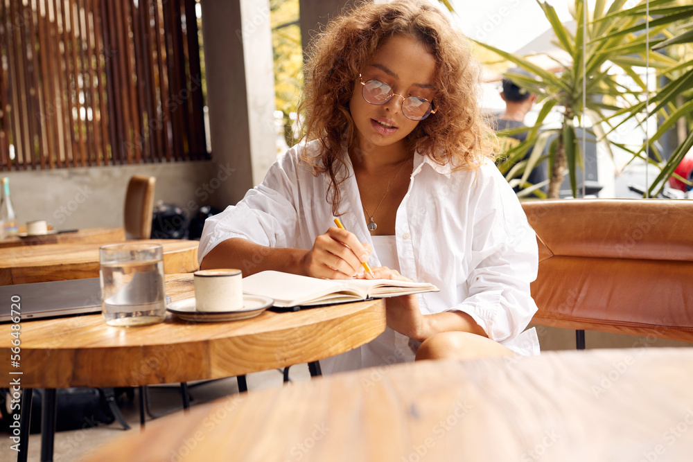 Freelancer At Cafe. Serious Woman In Glasses Writing Something in Notebook. Remote Job Or Education With Modern Digital Technologies For Comfortable Lifestyle In City Freelancer At Cafe. Serious Woman In Glasses Writing Something in Notebook. Remote Job Or Education With Modern Digital Technologies For Comfortable Lifestyle In City