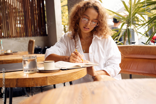 Freelancer At Cafe. Serious Woman In Glasses Writing Something in Notebook. Remote Job Or Education With Modern Digital Technologies For Comfortable Lifestyle In City