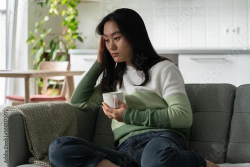 Photos Depressed weakened Asian woman with cup of coffee sits on sofa in living room of own home