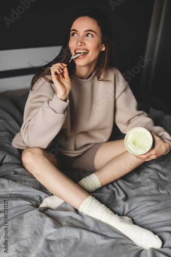 A young woman lies on her bed while eating a pint of pistachio ice cream with spoon. She is lick spoon with ice cream. Eating in bed. Happy beautiful woman resting in her comfortable bed at home. 
