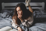 Charming caucasian lady lying in bed, reading a book, she look boring and tired.