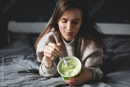 Tableau sur toile A young woman lies on her bed while eating a pint of pistachio ice cream with spoon