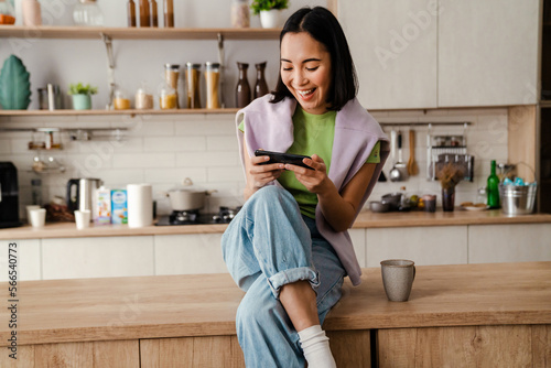 Excited asian woman playing online game on mobile phone while sitting in kitchen