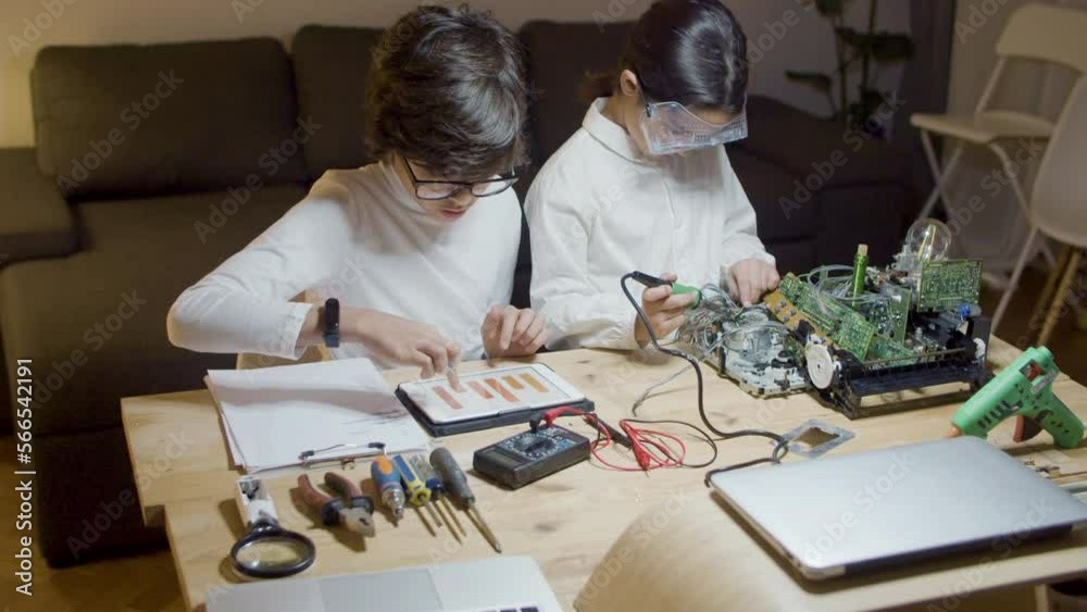 Two smart children sitting at desk and doing project work together at home. Girl soldering circuit board while boy in white shirt looking at bar chart on tablet and giving advice. Education concept Two smart children sitting at desk and doing project work together at home. Girl soldering circuit board while boy in white shirt looking at bar chart on tablet and giving advice. Education concept