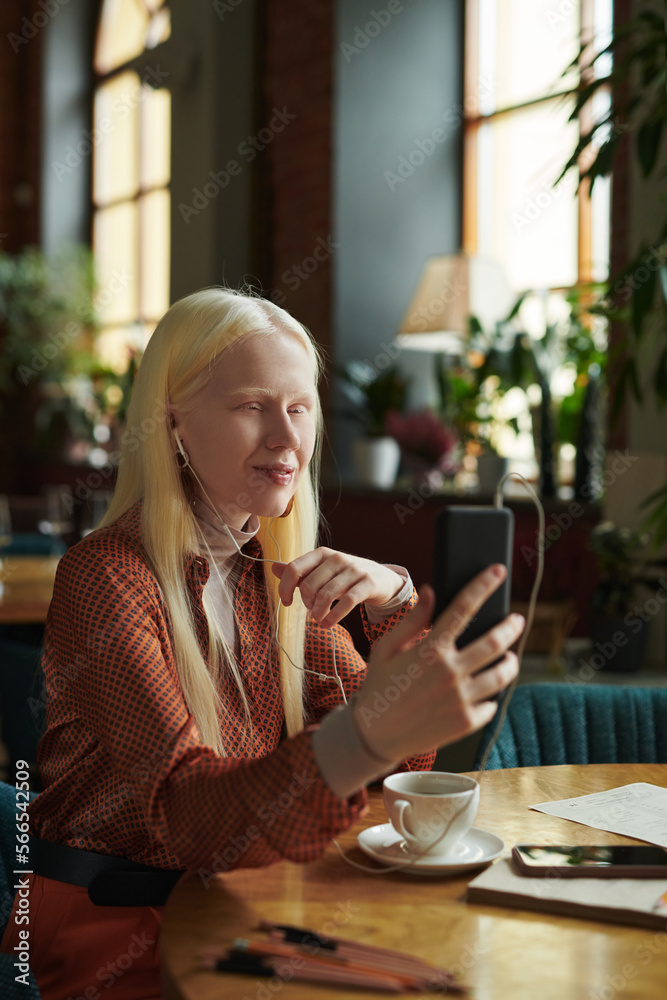 Young woman with long blond hair looking at smartphone screen during communication with her friend in video chat while sitting in cafe
