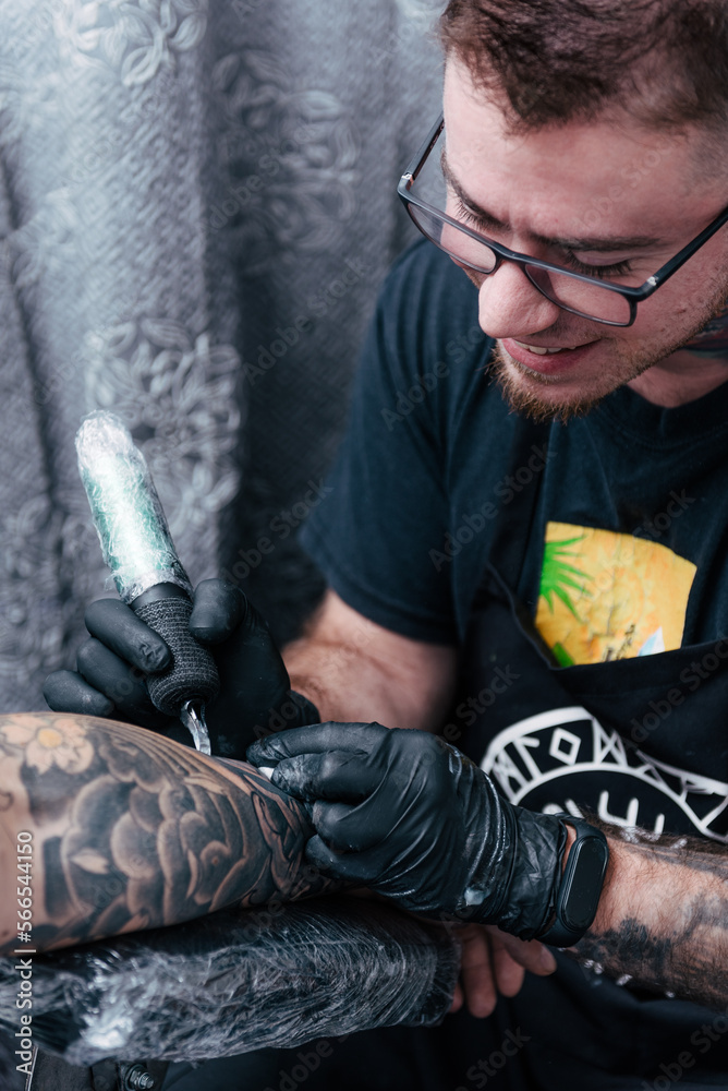 Obraz premium Detail of the tattooist's hand with a black glove holding the tattoo machine while loading ink into the needle while making a tattoo. Vertical