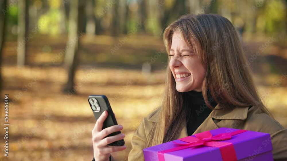 Side view of young, beautiful woman walking in park in autumn. Attractive, stylish female standing, holding present, gift box, using smartphone. Concept of modern lifestyle. Side view of young, beautiful woman walking in park in autumn. Attractive, stylish female standing, holding present, gift box, using smartphone. Concept of modern lifestyle.