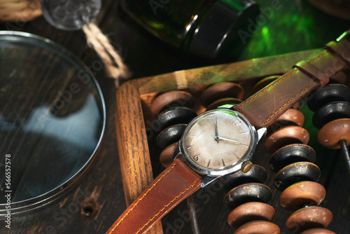 Vintage men's Omega dress watch with white rustic dial and brown leather strap laying on the wooden abacus near to the glass bottle and magnifier on the brown wooden surface in a daylight.