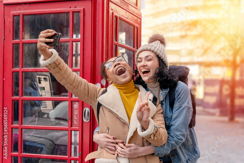 Photography two friend, girlfriend and women using a mobile phone, camera and taking selfie  against a red phonebox in the city of England