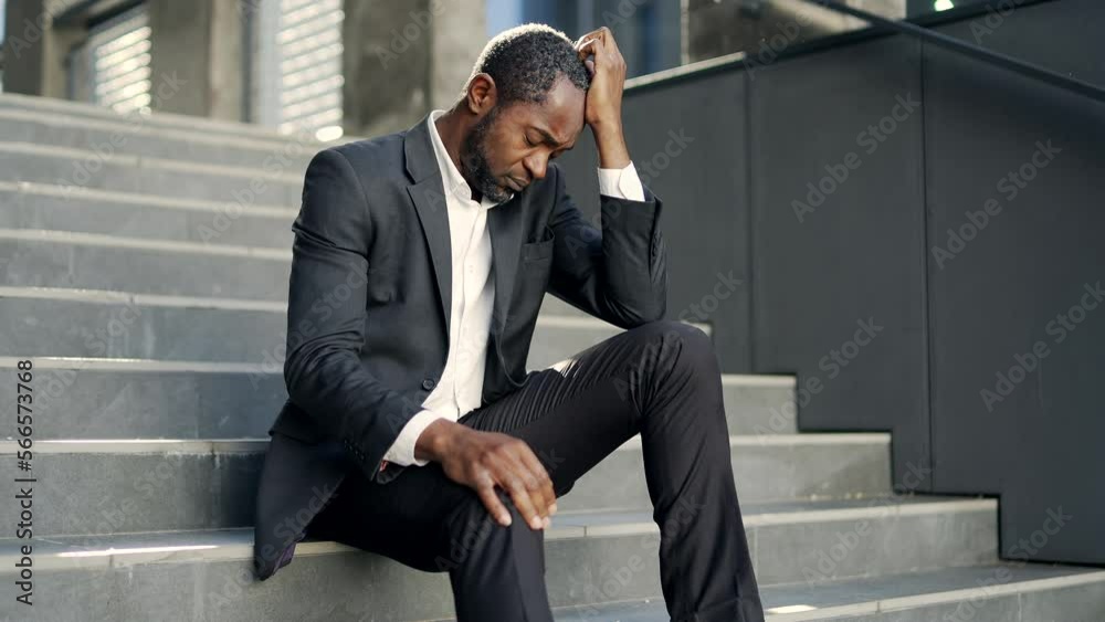 Sad african american mature man sitting outside on the steps of an office building. Middle-aged male in a formal suit holds his hand on his head. Вusinessman went bankrupt or an employee lost his job Sad african american mature man sitting outside on the steps of an office building. Middle-aged male in a formal suit holds his hand on his head. Вusinessman went bankrupt or an employee lost his job