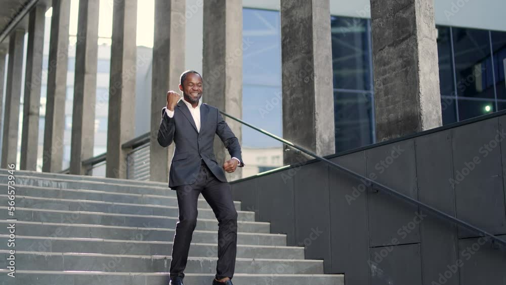Happy excited african american businessman celebrating success joyfully running down stairs of office building Middle-aged man in a formal suit is dancing, holding his hands up in a triumphant gesture Happy excited african american businessman celebrating success joyfully running down stairs of office building Middle-aged man in a formal suit is dancing, holding his hands up in a triumphant gesture