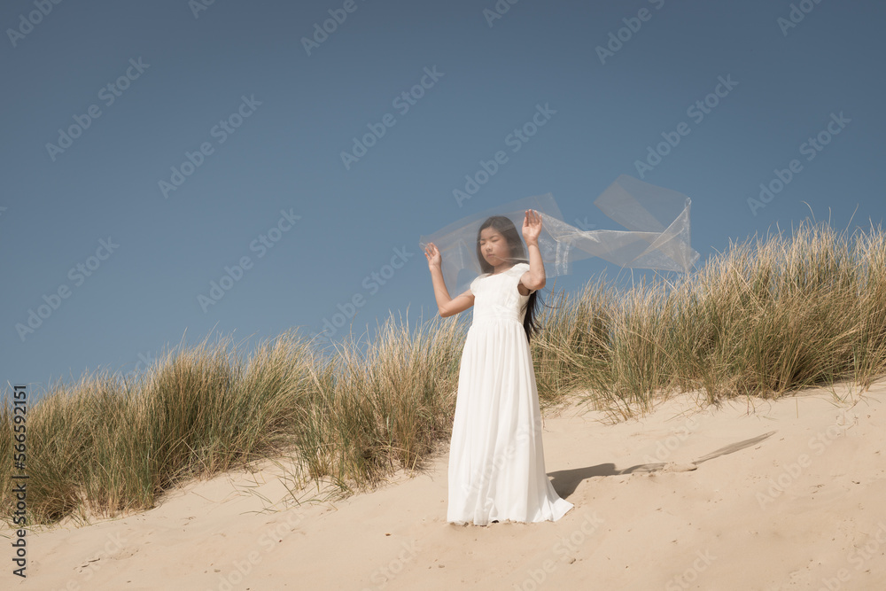 asian girl in white long classic dress standing in sunlight in dunes near ocean holding veil in the wind asian girl in white long classic dress standing in sunlight in dunes near ocean holding veil in the wind