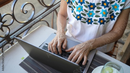 Closeup looking down on a mature old womans hands using a laptop computer with a drink on a table. Concept of digital nomad traveling the world.
