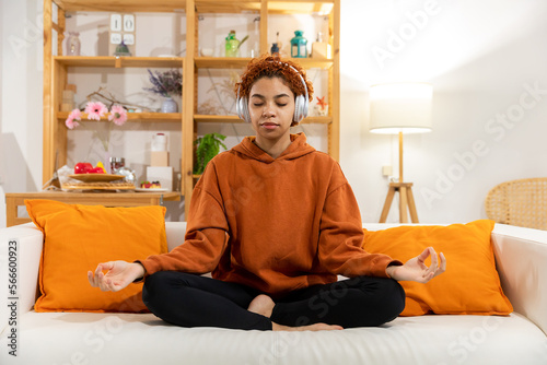 Yoga mindfulness meditation. Young healthy african girl practicing yoga at home. Woman sitting in lotus pose on couch meditating smiling relaxing indoor. Girl doing breathing practice. Yoga at home