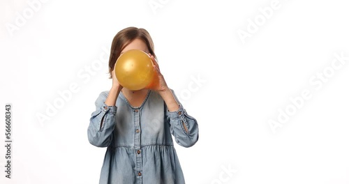 Pretty little girl in casual denim dress blowing, inflate yellow balloon on white background, studio