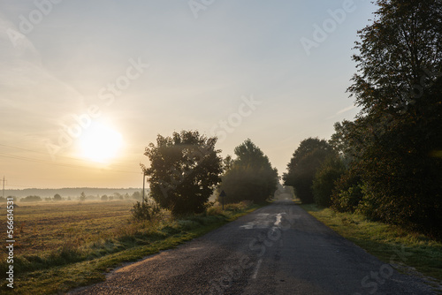 Road in rural landscape with fields both sides. Early morning with mist and fog. Glow from rising sun.