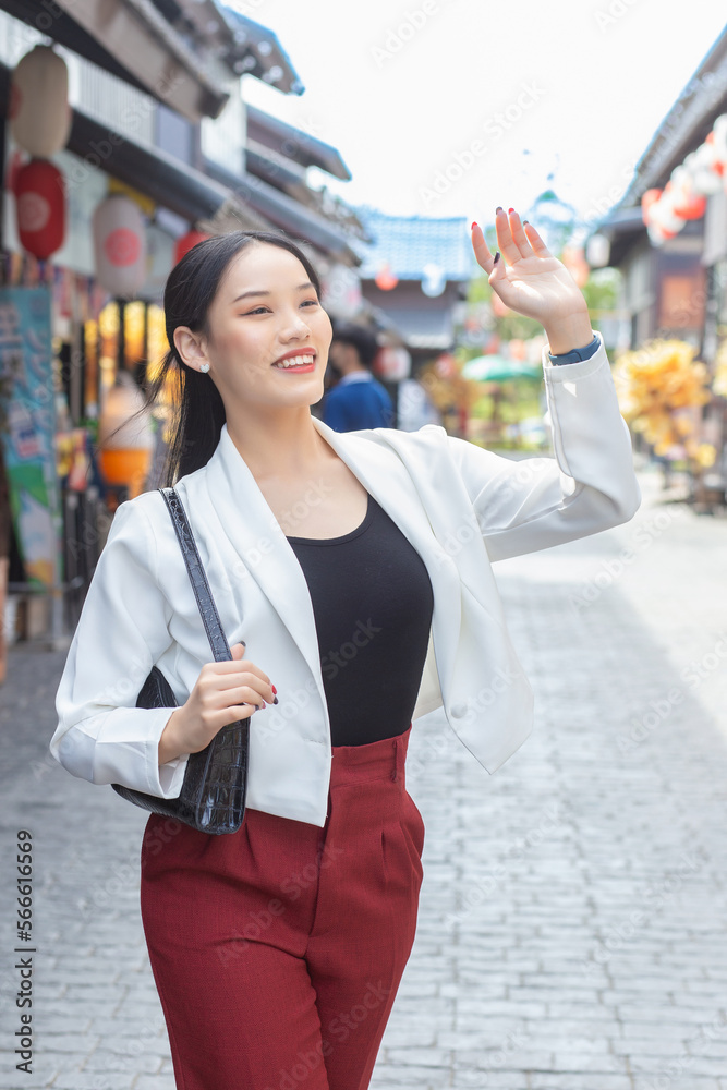 Fototapeta premium Young beautiful Asian business woman wear white suit and red pants smiling confidently,carry bag while walking in the city on a sunny day.