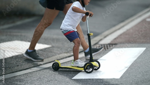 Child crossing street with scooter on zebra lines. Parent and kid on crosswalk in sunny day. Active small boy rides trandportation crosses sidewalk