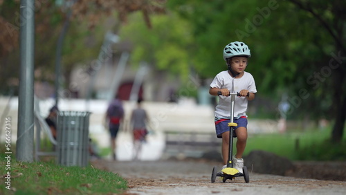 Carefree child riding scooter at park. One male kid wearing helmet rides three wheeled toy scooter outdoors. Sportive childhood concept in slow motion