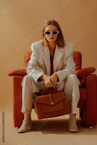 Fashionable confident woman wearing elegant white suit, sunglasses, ankle boots, holding classic brown leather bag, sitting in armchair, posing on beige background. Full-length studio fashion portrait