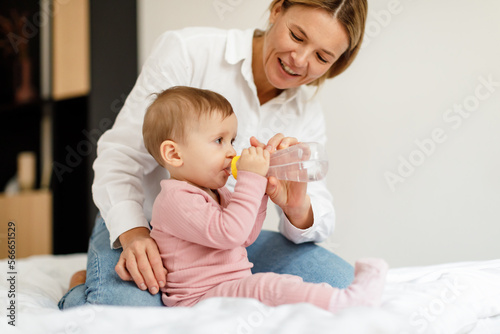 Baby hydration. Caring mother giving water bottle to her infant child, sitting on bed at home, copy space