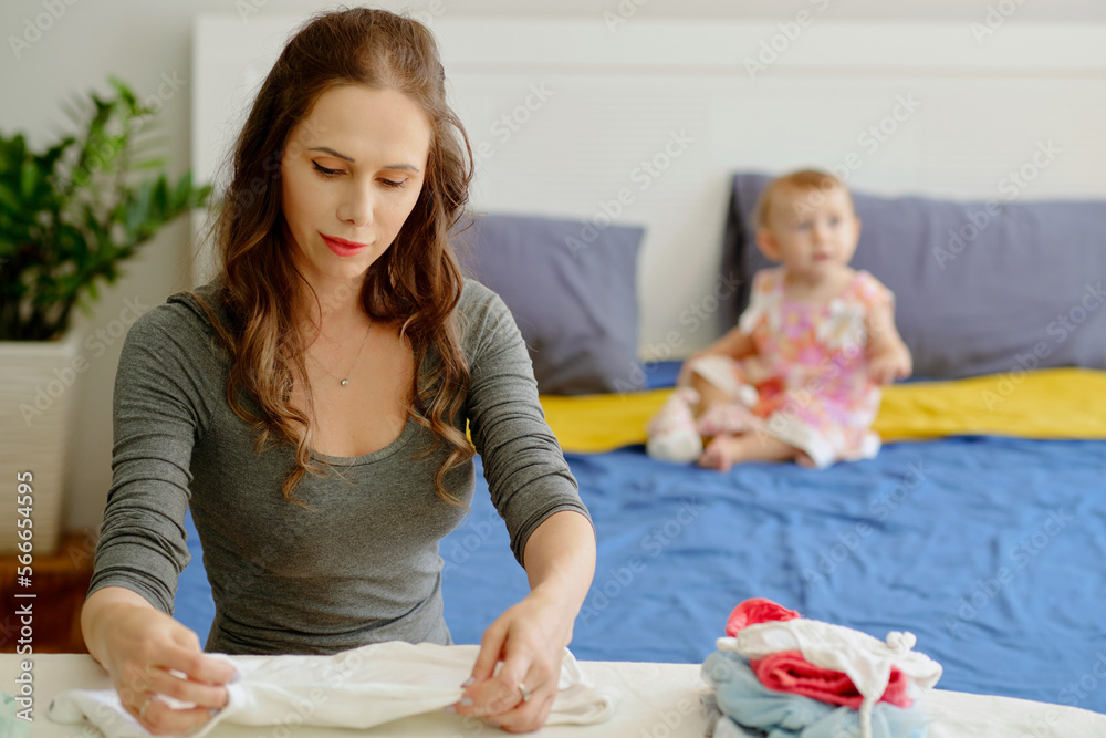 Young woman ironing and folding clothes when her daugher playing in background Young woman ironing and folding clothes when her daugher playing in background